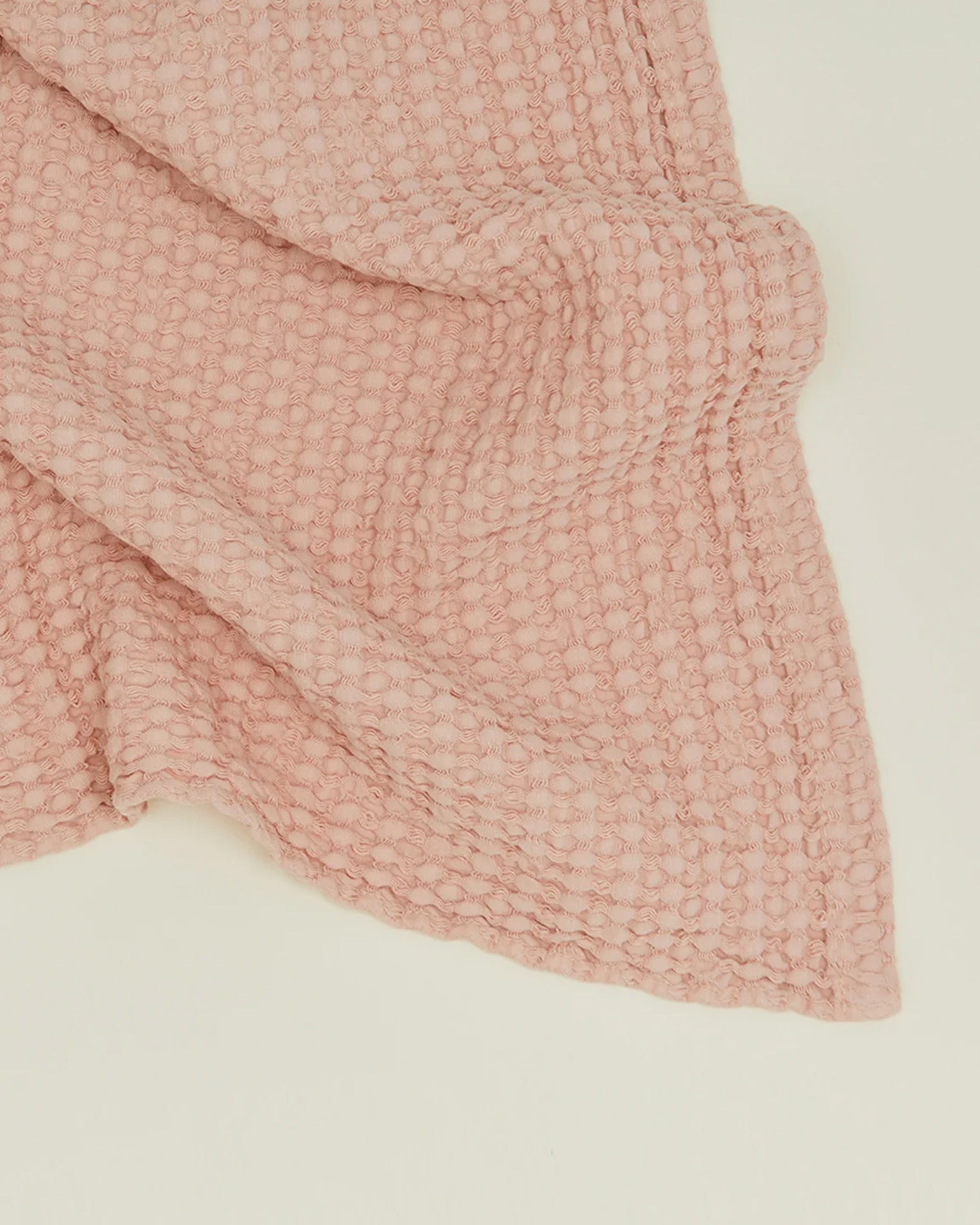The Hawkins New York Simple Waffle Hand Towel in Blush features a textured, waffle-like pattern and light pink hue, crafted in Portugal to add elegance and charm to any space. Draped on a plain, light background.