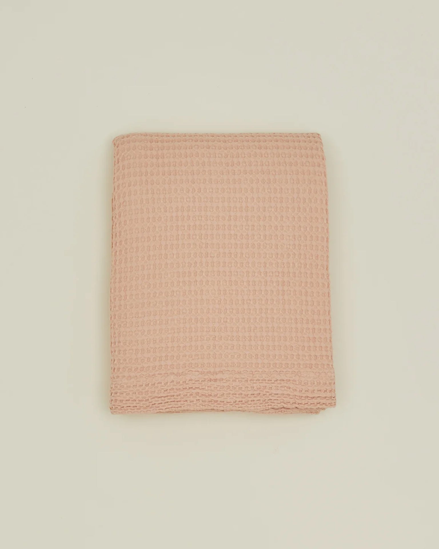 A folded, blush waffle-textured towel rests on a plain off-white background, evoking the delicate weave of the Hawkins New York Simple Waffle Shower Curtain.