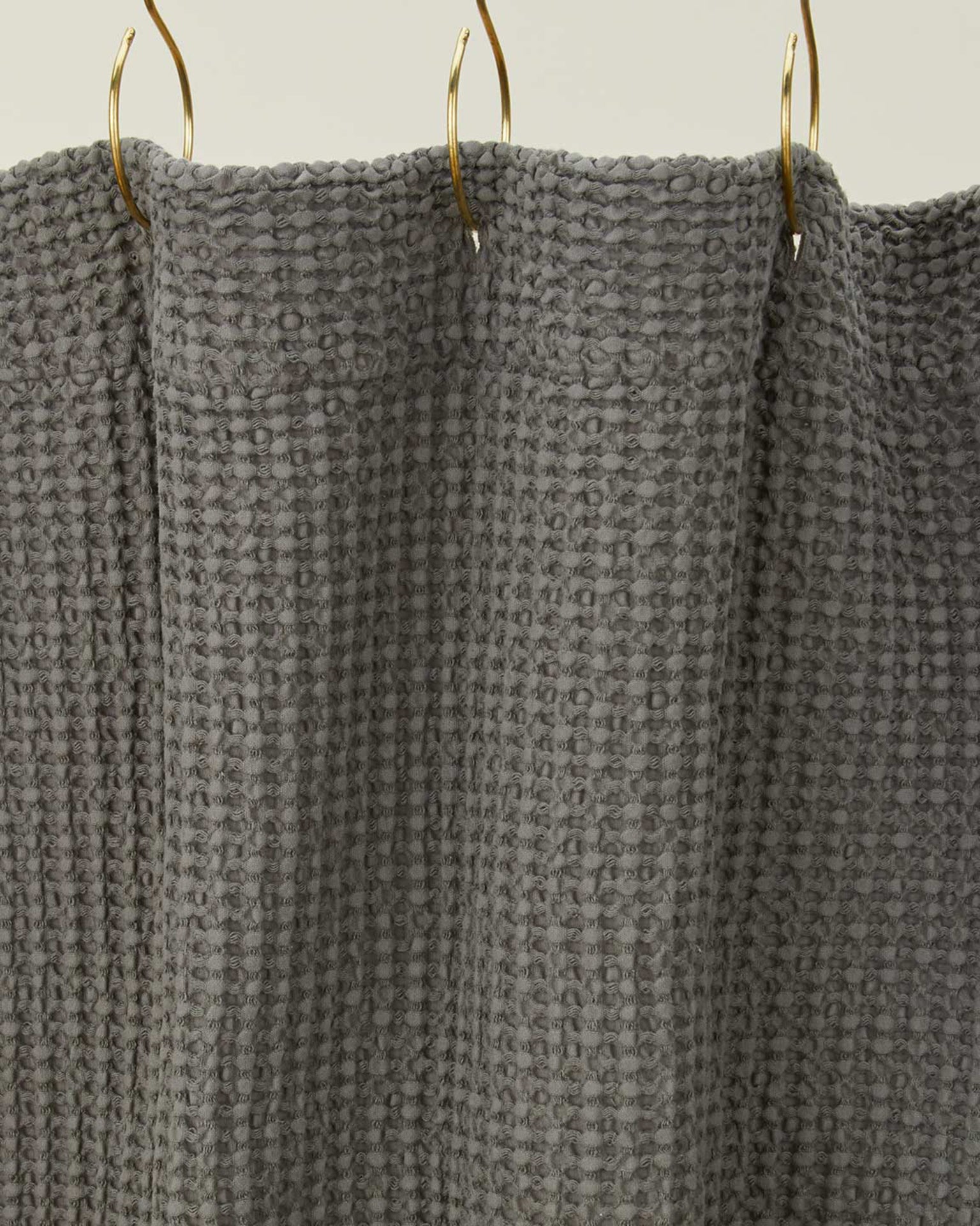 A close-up of the Simple Waffle Shower Curtain in Dark Grey by Hawkins New York, made of cotton with a textured waffle pattern, hanging from gold hooks.