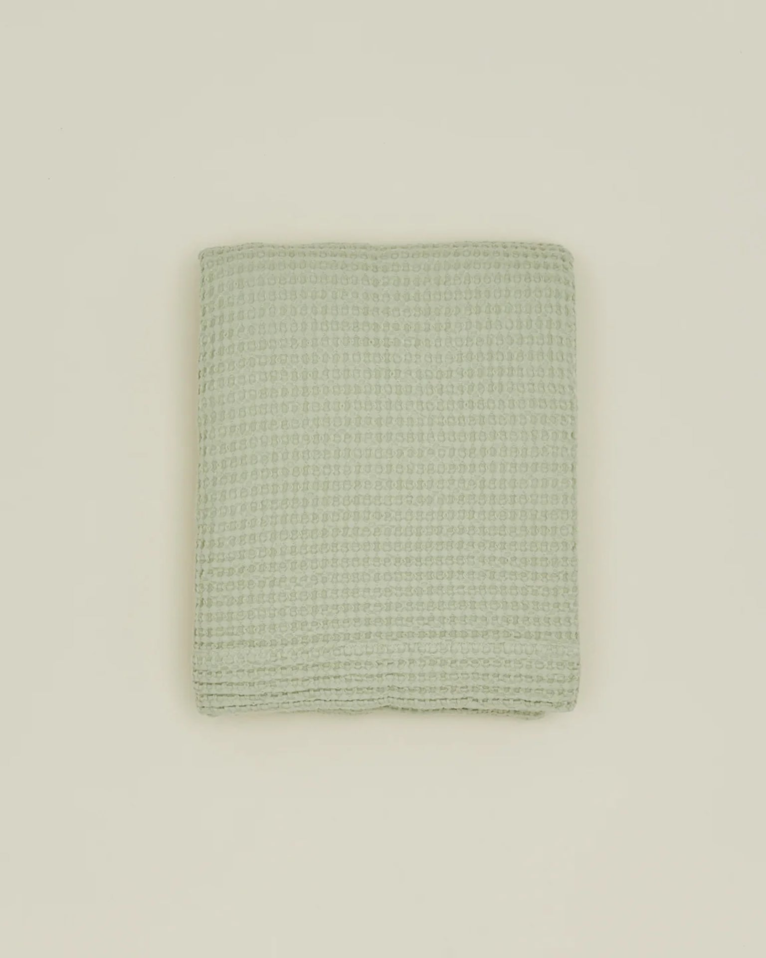A folded Simple Waffle Shower Curtain in Sage by Hawkins New York, featuring a waffle texture, is centered on a plain beige background.