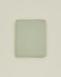A folded Simple Waffle Shower Curtain in Sage by Hawkins New York, featuring a waffle texture, is centered on a plain beige background.