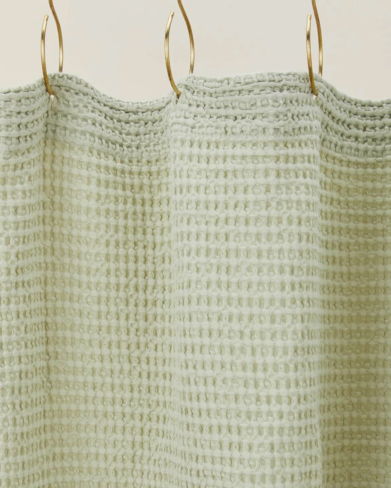 The Simple Waffle Shower Curtain in Sage by Hawkins New York features a light green cotton waffle texture, shown on gold hooks against a neutral backdrop, showcasing the brand’s timeless style.