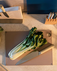 A bunch of bok choy and a kitchen knife rest on The (grippy) reBoard® | Taffy cutting board by Material, with knife blocks and utensils in the background.