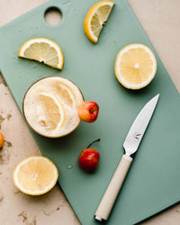 The (mini) reBoard® | Moss by Material, made from sustainable materials, showcases a halved lemon, lemon slices, cherries, a small knife, and a glass of frothy lemon drink.