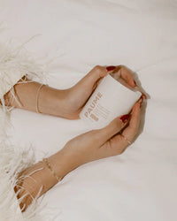 A person with manicured nails and gold jewelry holds a white PAUME Overnight Hand & Foot Hydration Mask jar against a soft, white, textured background.