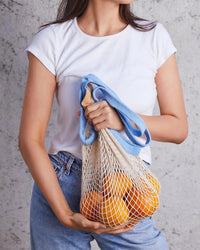 A person holds PILLOWPIAs The French Market Bag 2Tone In Wheat w/ French Blue, filled with oranges, against a gray background.