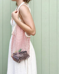 A person in a white sleeveless dress carries PILLOWPIAs The French Market Bag No.2 in Ballet Pink, filled with items, while standing before a light green wooden wall.