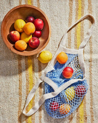 A wooden bowl of assorted fruit sits beside PILLOWPIA’s The French Market Bag No.2 in French Blue, filled with similar fruit, on a striped fabric surface.