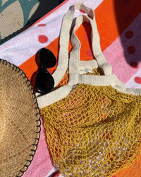 A straw hat, black sunglasses, and PILLOWPIAs French Market Bag No.2 in Goldenrod are arranged on a pink and orange striped towel in sunlight.