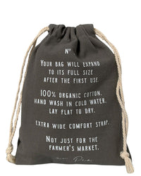 The French Market Bag No.2 in Iron by PILLOWPIA is a black drawstring bag with white care instructions, made from 100% organic cotton, featuring an extra wide comfort strap and recommended cold hand washing.
