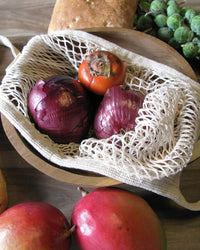 The French Market Bag No.2 in Natural by PILLOWPIA holds two red onions and a persimmon in a wooden bowl, with bread, Brussels sprouts, and fruit displayed nearby on a wooden surface.