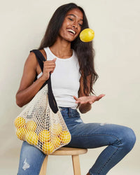 A woman sits on a stool, holding PILLOWPIA’s French Market Bag No.2 in Natural with Monochrome Black, filled with yellow apples, and tosses one apple in the air.