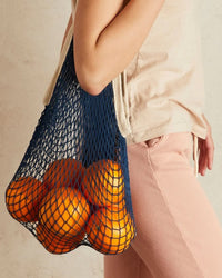 A person holds PILLOWPIAs The French Market Bag No.2 in Navy, filled with oranges, while wearing a beige shirt and pink pants against a light-colored wall.