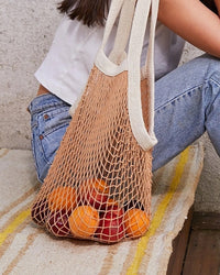 A person sits on a rug with the PILLOWPIA French Market Bag No.2 in Wheat, filled with oranges and apples, resting on the floor.