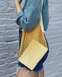 A person in a blue long-sleeve shirt and denim shorts carries PILLOWPIA’s The French Market Bag No.2 in Yolk with a square item inside, standing against a white brick wall.