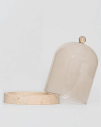 Poured New York’s Cloche + Stone in Oyster showcases a glass dome with round handle, stylishly angled on a circular beige stone base—an exquisite luxury accent for modern home décor.