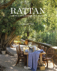 A rustic outdoor dining table with Random House Rattan furniture sits under large trees amid greenery. A lantern hangs above, and the table is set with books, dishes, and glassware.