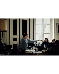 Four people sit around a table with Spilled Natural Winemakers Stories & Recipes by Random House, enjoying wine, as one person stands to hand an item to someone near a window with sheer curtains.