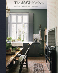 The deVOL Kitchen by Random House features dark cabinets, a wooden dining table, houseplants, and natural light with a black stove on herringbone brick—ideal inspiration for unique and timeless kitchen design.