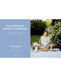 A woman in a light dress sets food on an outdoor table by flowers. On the left is The Wishbone Kitchen Cookbook, published by Random House, featuring dinner party recipes by Meredith Hayden and contributor credits.