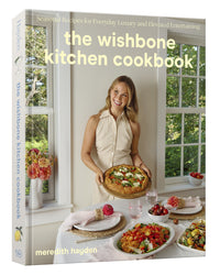 A woman stands by a set dining table holding a pizza in front of a window; featured is The Wishbone Kitchen Cookbook by Random House.