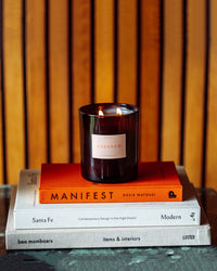A lit Le Grand Dresden coconut soy wax candle by ROEN sits atop three stacked books, the top one being Manifest by Roxie Nafousi, with wood paneling in the background.