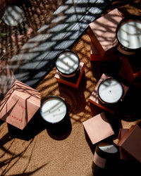 Le Grand Marmont luxury scented candles by ROEN, in dark glass jars, sit on a surface with pink boxes as sunlight and leaf shadows play across the scene.