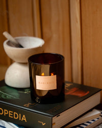 A lit ROEN Le Grand Ojai Nuit candle, crafted from coconut soy wax, rests atop a stack of books beside a white mortar and pestle, with wood paneling in the background.