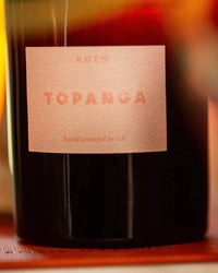 A close-up of ROEN’s Le Grand Topanga candle shows a dark glass bottle with a pink textured label; this vegan soy wax candle offers a fresh, earthy scent and is hand-poured in California.