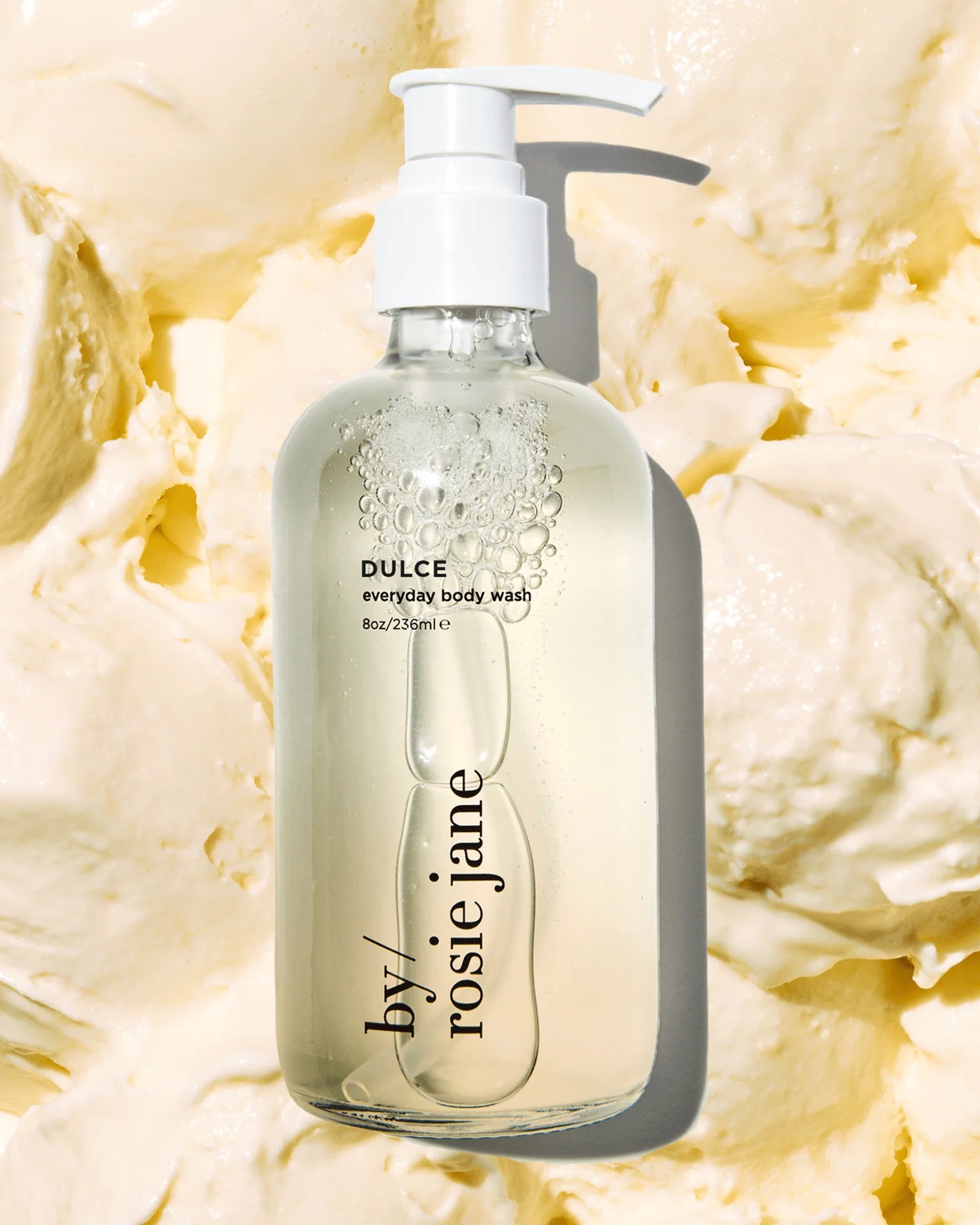 A clear pump bottle of Rosie Jane Dulce Everyday Body Wash sits on a creamy, textured off-white background that looks like swirled lotion or whipped cream.