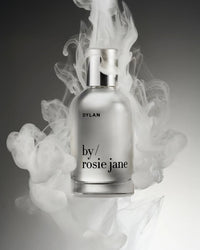 A clear Eau De Parfume in Dylan bottle by Rosie Jane is enveloped in swirling white smoke on a gray background, capturing the unisex fragrances signature white musk essence.