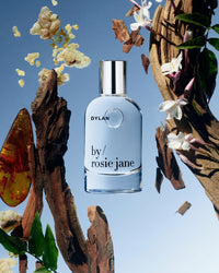 A bottle of Rosie Jane Eau De Parfum in Dylan is shown among floating wood, flowers, amber, and leaves against a blue background, evoking its signature white musk scent.