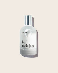 A clear glass bottle of Eau De Parfume in Dylan by Rosie Jane, with a silver cap, displayed against a light beige background.