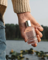 Wearing a beige knit sweater, a person holds a clear bottle of Rosie Jane Eau De Parfume in Lake—a perfect summer scent—beside a lake with water lilies and trees in the background.