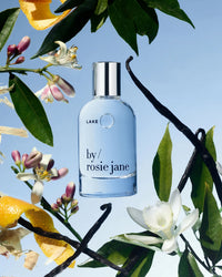 Rosie Jane Eau De Parfume in Lake is displayed among leaves, flowers, and orange peels with a blue sky backdrop.