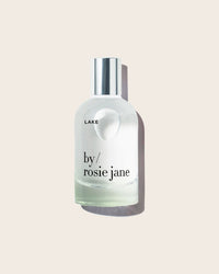 A clear glass bottle of Eau De Parfume in Lake by Rosie Jane, topped with a silver cap and placed on a light beige background, suggests a fresh and summery scent.