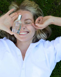 A woman in a white shirt smiles with her eyes closed, lying on grass and holding Rosie Jane’s Eau De Parfume in Leila Lou close to her face.