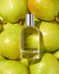 A bottle of Eau De Parfume in Leila Lou by Rosie Jane, a fresh floral fragrance, rests on green apples with sparkling water droplets.