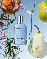 A bottle of Eau De Parfume in Leila Lou by Rosie Jane sits on a blue background, surrounded by jasmine flowers, a peeled orange, a green lemon, and sliced pear.