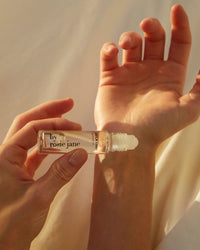 A hand applies Rosie Jane’s Perfume Oil in Leila Lou to another’s wrist, as soft sunlight highlights the gesture and evokes a delicate floral scent.