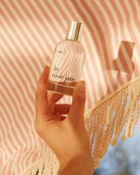 A hand holds a clear Eau De Parfume in Missy by Rosie Jane in front of a pink and white striped fringed fabric.