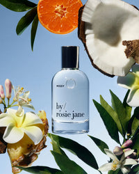 A bottle of Rosie Jane’s Eau De Parfume in Missy sits on a blue background, surrounded by toasted coconut, an orange slice, flowers, green leaves, and pineapple.