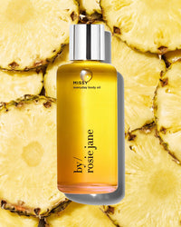 A bottle of Missy Everyday Body Oil by Rosie Jane stands upright on fresh pineapple slices. The hydrating oil comes in a yellow bottle with a silver cap and minimalist black text.