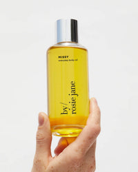 A hand holds a clear bottle of Missy Everyday Body Oil by Rosie Jane, featuring a silver cap, shown against a plain white background.