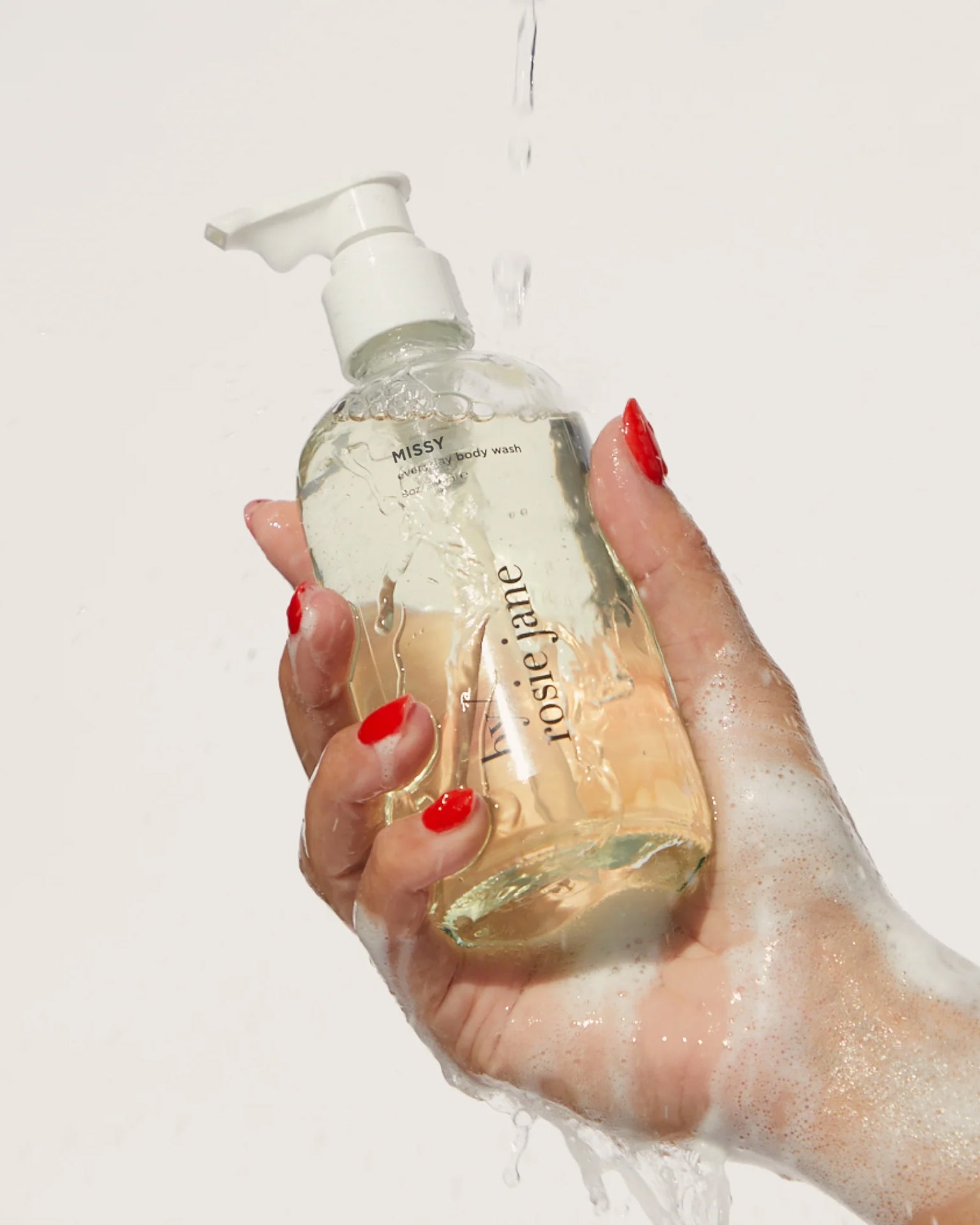 A hand with red-painted nails holds a clear pump bottle labeled Rosie Jane Missy Everyday Body Wash, a creamy hydrating cleanser covered in lather and water droplets against a plain light background.