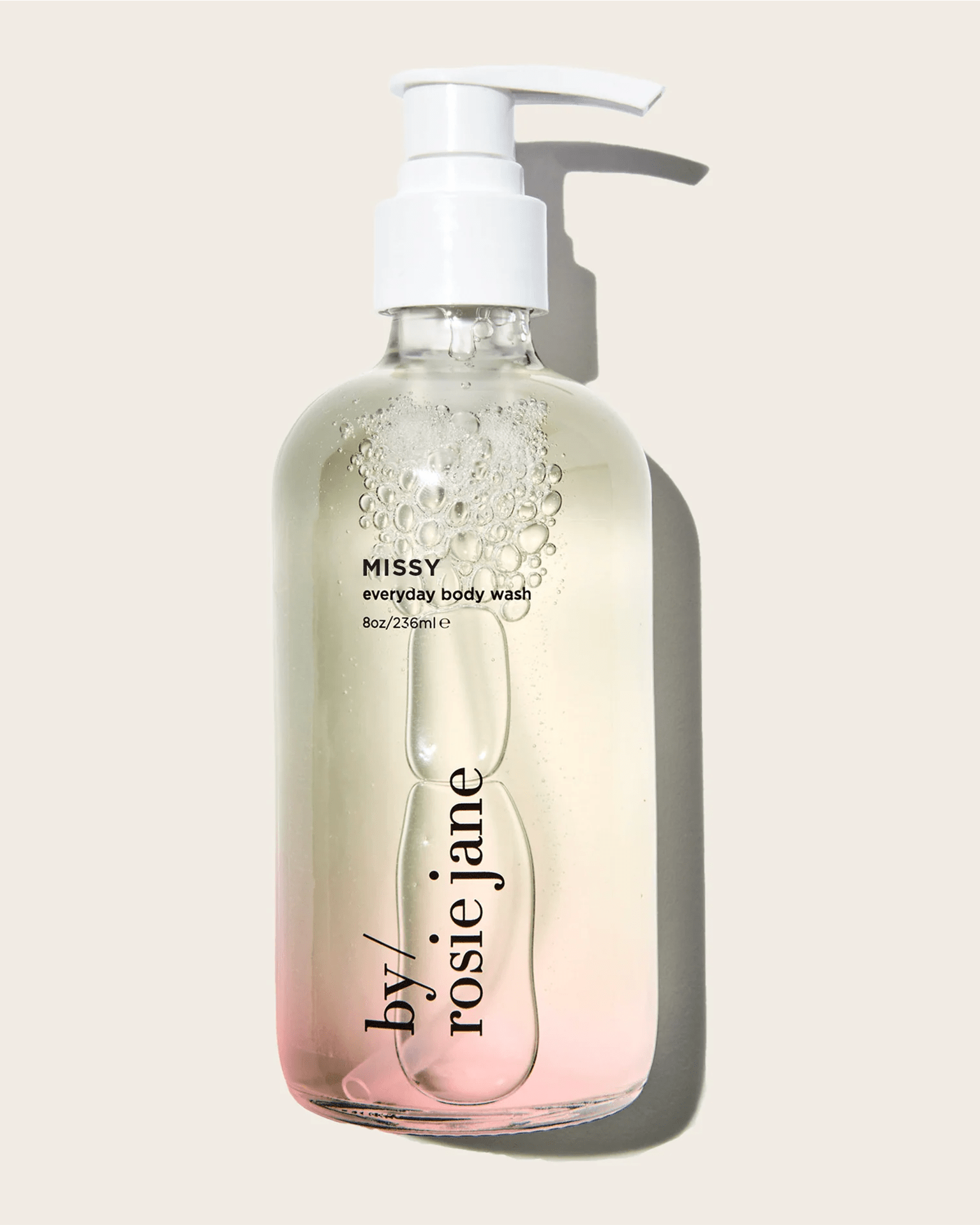 A clear pump bottle of Rosie Jane Missy Everyday Body Wash, 8oz/236ml, with liquid fading from clear to light pink, sits against a beige background—an everyday hydrating cleanser for skin kissed with the DULCE scent.