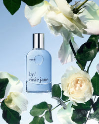 The Rosie Jane Eau De Parfume in Rosie is showcased among dewy white roses, capturing a soft skin scent vibe against a light blue backdrop.