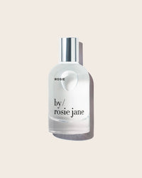 A clear glass Eau De Parfume in Rosie by Rosie Jane, with a silver cap and labeled ROSIE, sits on a light beige background—ideal for lovers of soft white musk fragrances or delicate skin scents.