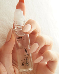 A hand with long, nude nails holds a clear glass bottle of Rosie Jane Perfume Oil in Rosie, featuring notes of rose essential oil and white musk, shown with a rollerball applicator against a soft, neutral background.