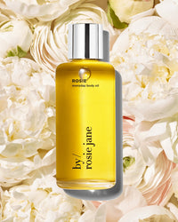 A bottle of Rosie Jane’s Rosie Everyday Body Oil with a silver cap stands upright among cream peony flowers, its golden yellow hydrating formula contrasting beautifully with the soft floral backdrop.